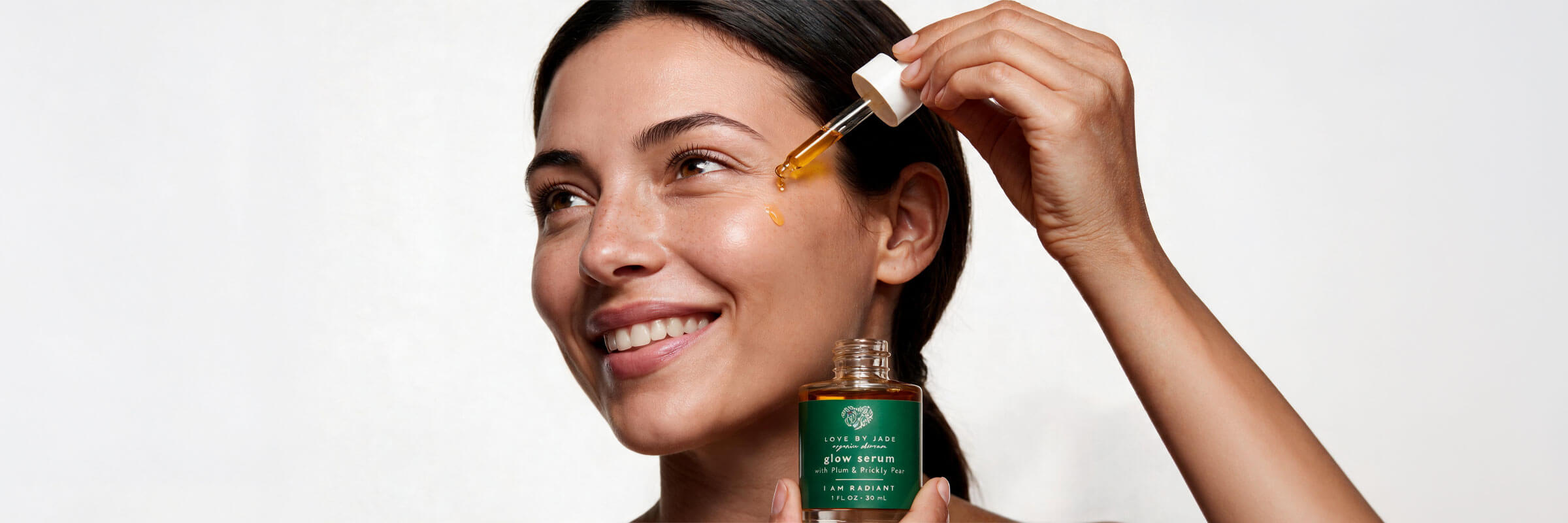 Woman smiling as she holds the Love by Jade Glow Face Serum dropper beside her face