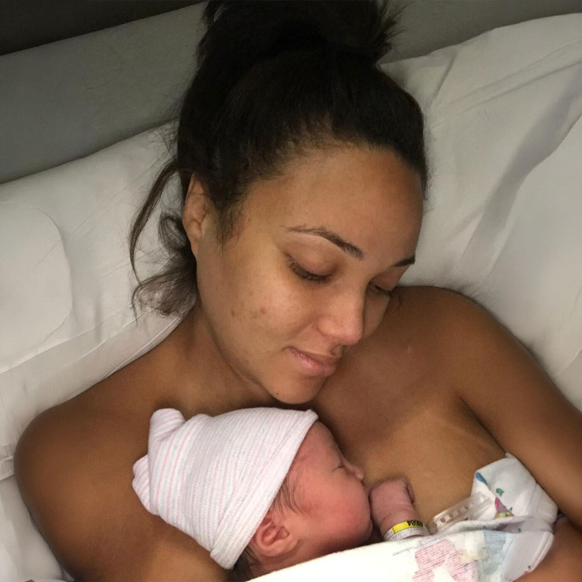 A close-up lifestyle shot of a Jade breastfeeding a newborn baby in a hospital bed. The woman has her hair in a bun and is looking down at the baby, who is wearing a white hat and lying on a white blanket.
