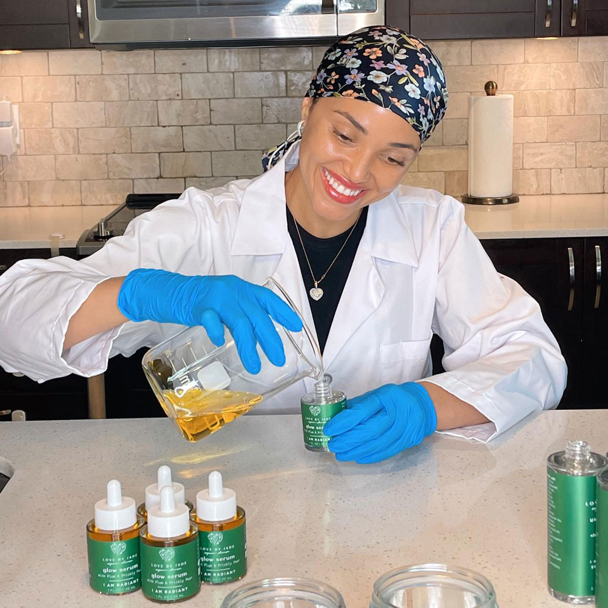 Jade wearing a white lab coat, blue gloves, and a floral headscarf pours a golden liquid from a glass beaker into a small green bottle on a kitchen counter. Several small amber dropper bottles with green labels are lined up in front of her.