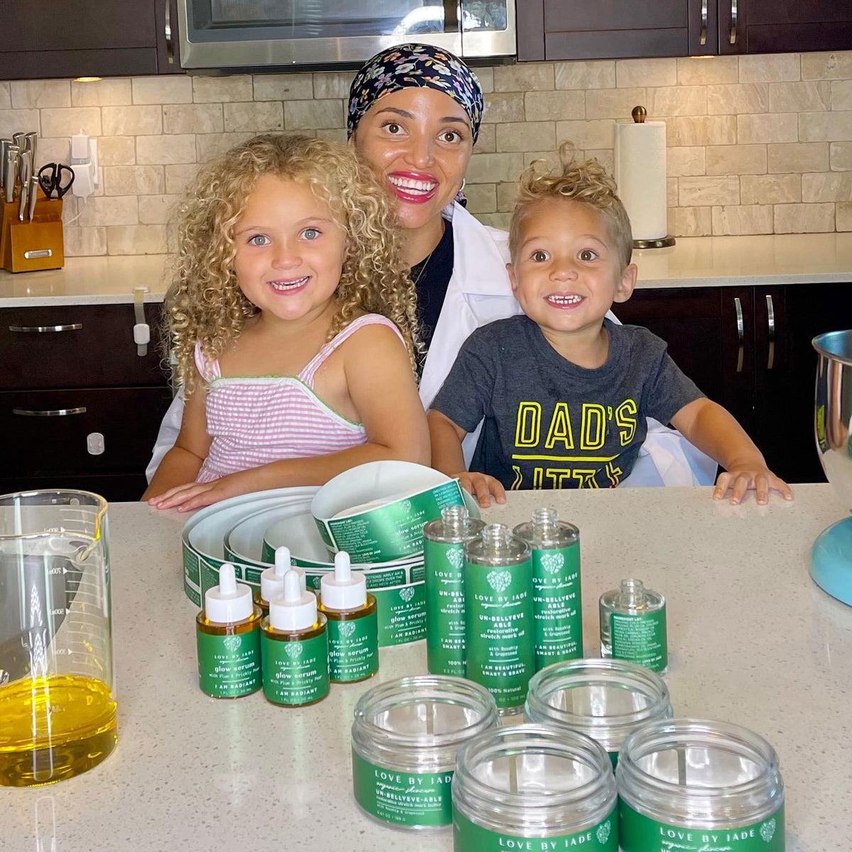 Jade and her two children standing in a kitchen behind a counter filled with green-labeled skincare or cosmetic containers, including dropper bottles and jars. The products appear to be the items being sold, but the scene includes people and a real kitchen environment, so subject_type is Lifestyle rather than Product/Packaging.